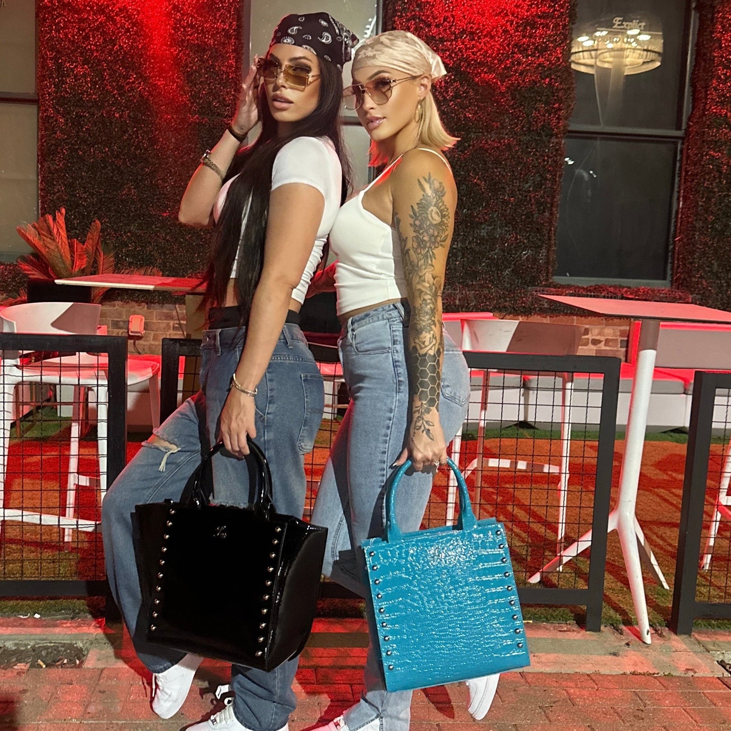 Two women posing outside a building with a neon sign, holding the Laurice Noire Anastacia Gold and Anastacia 2 handbags.