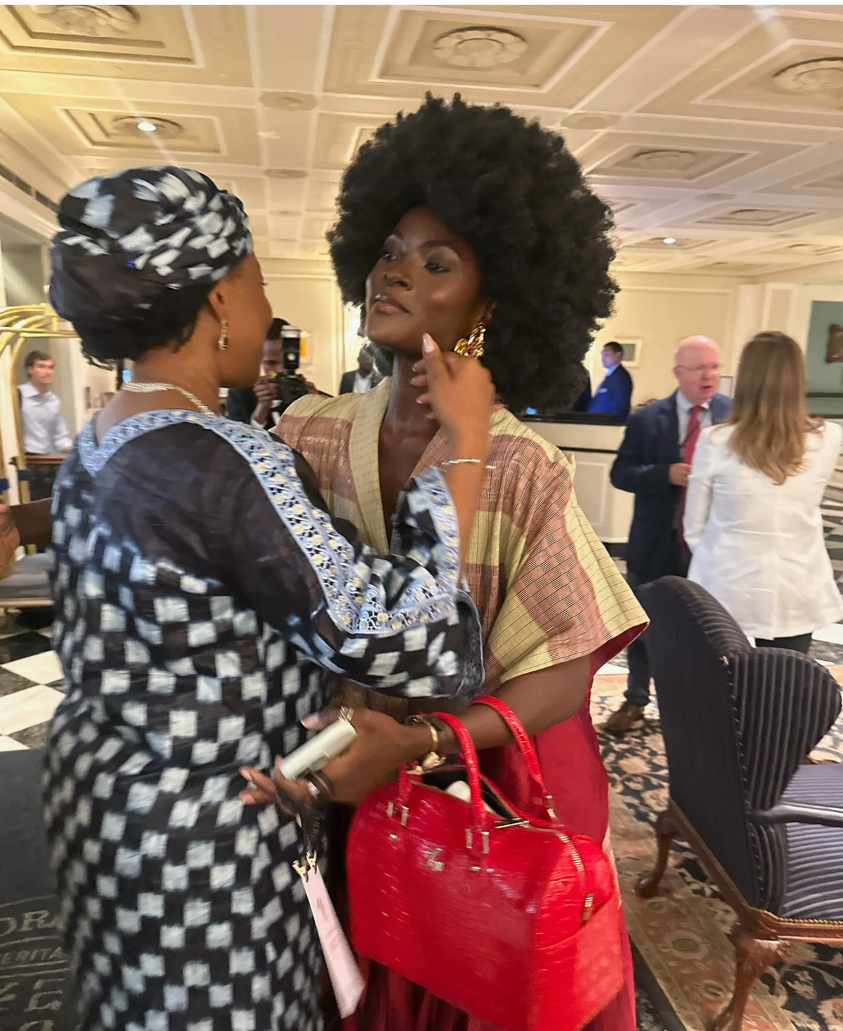 Two women interacting in a formal setting with a red handbag.
