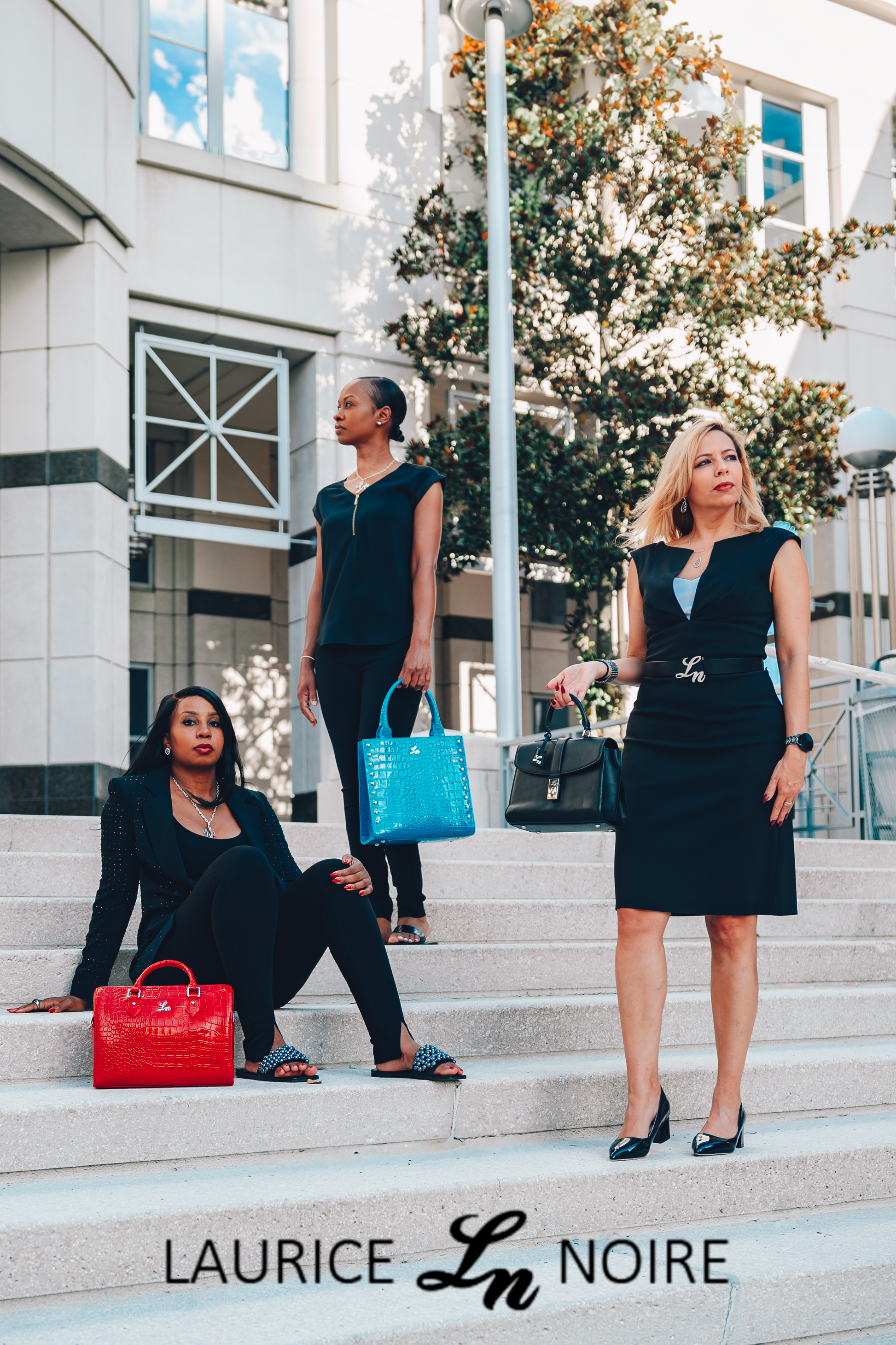 Three women with professional attire posing with Laurice Noire handbags, belts, and jewlery.