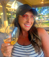 Woman in sunglasses holding a glass of wine, wearing a black cap and striped tank top, enjoying a sunny day.