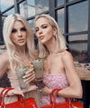 Two women holding cocktails, one in a pink dress and the other in a beige dress, enjoying a sunny day outdoors.
