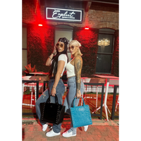 Two women standing outside a bar with neon signs, one holding a black handbag and the other a blue handbag.
