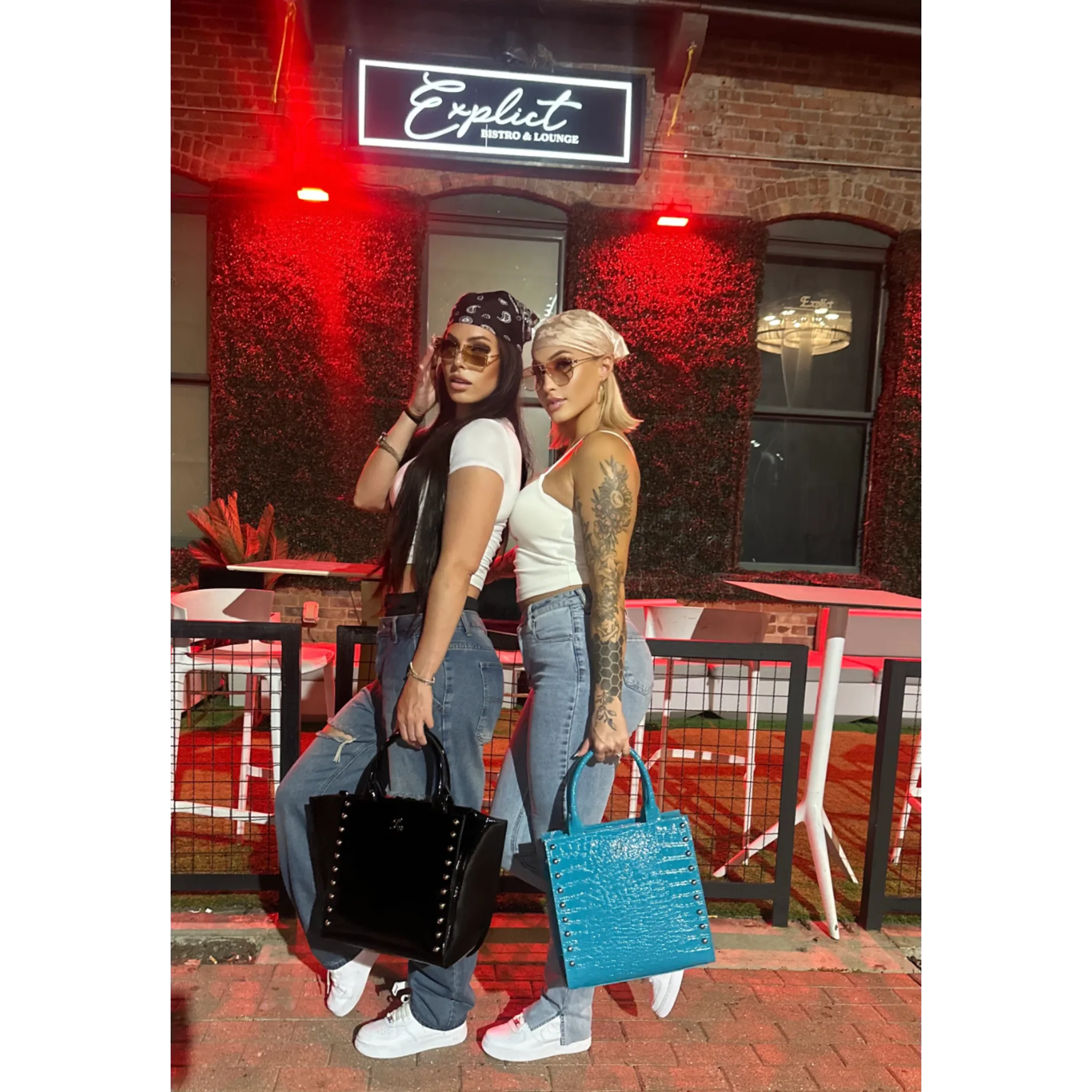 Two women standing outside a bar with neon signs, one holding a black handbag and the other a blue handbag.