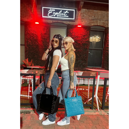 Two women standing outside a bar with neon signs, one holding a black handbag and the other a blue handbag.