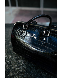 A glossy black crocodile-embossed handbag with silver hardware and a silver script logo reading 'Ln', resting on a dark stone surface.