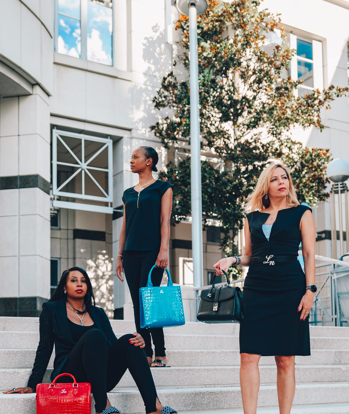 Three women in professional attire standing on steps outside a building with trees and a clear sky.