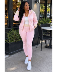 A woman modeling a light pink two-piece tracksuit with pearl-beaded trim on a sidewalk in front of a storefront. The set includes a zip-up hoodie and matching jogger pants, styled with a white tank top and white sneakers.
