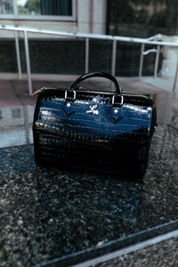 Black genuine patent croc leather handbag on a granite surface, showcasing gold hardware and logo detail.