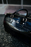Close-up of a black patent croc leather duffle bag with gold hardware and stitched initials.