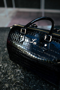 Close-up of a black patent croc leather duffle bag with gold hardware and stitched initials.