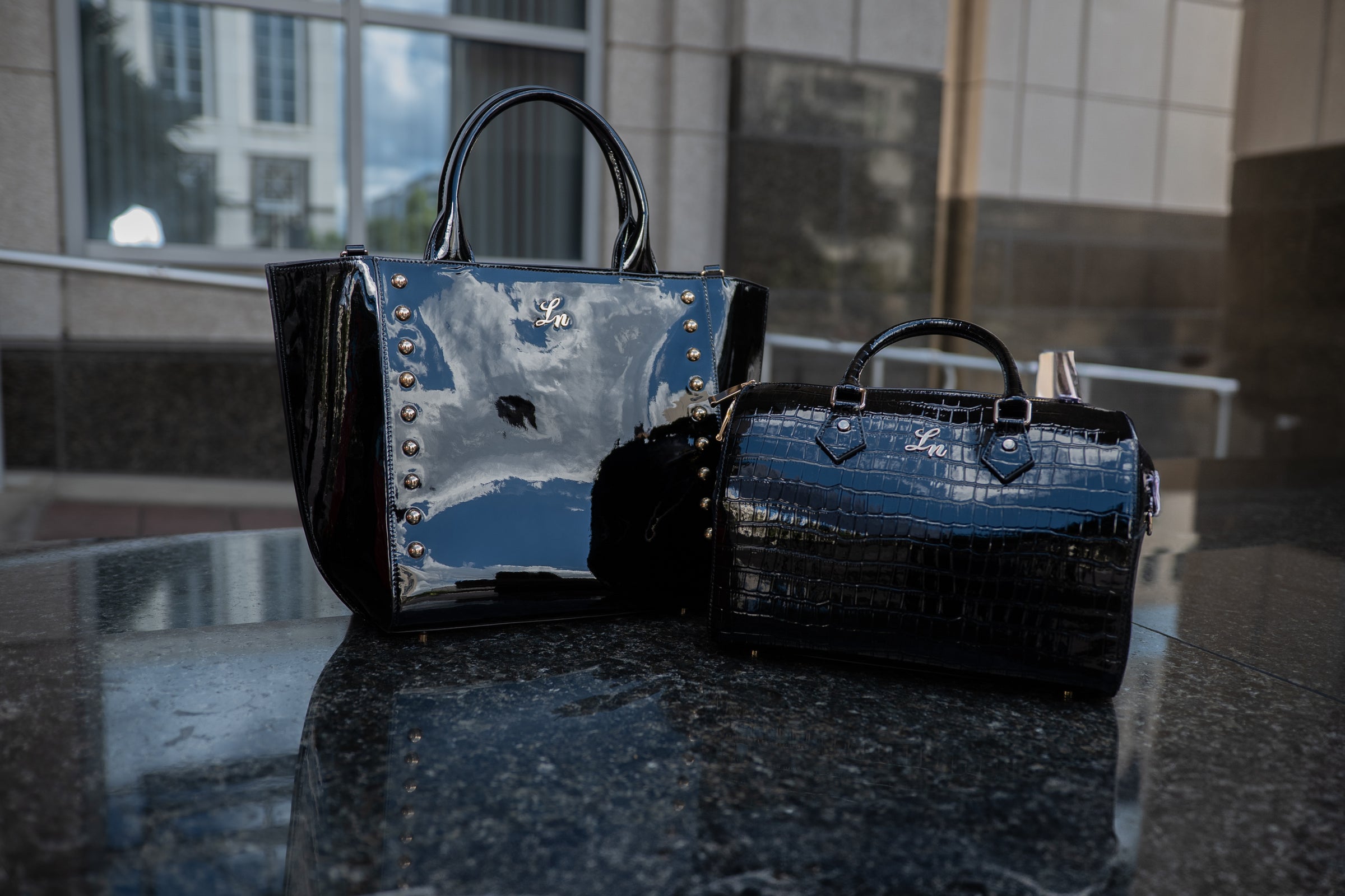 Two black patent leather handbags displayed on a reflective surface with gold studs and croc texture.