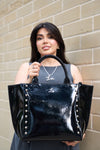 Woman holding a black patent leather handbag with studded details against a beige brick wall.