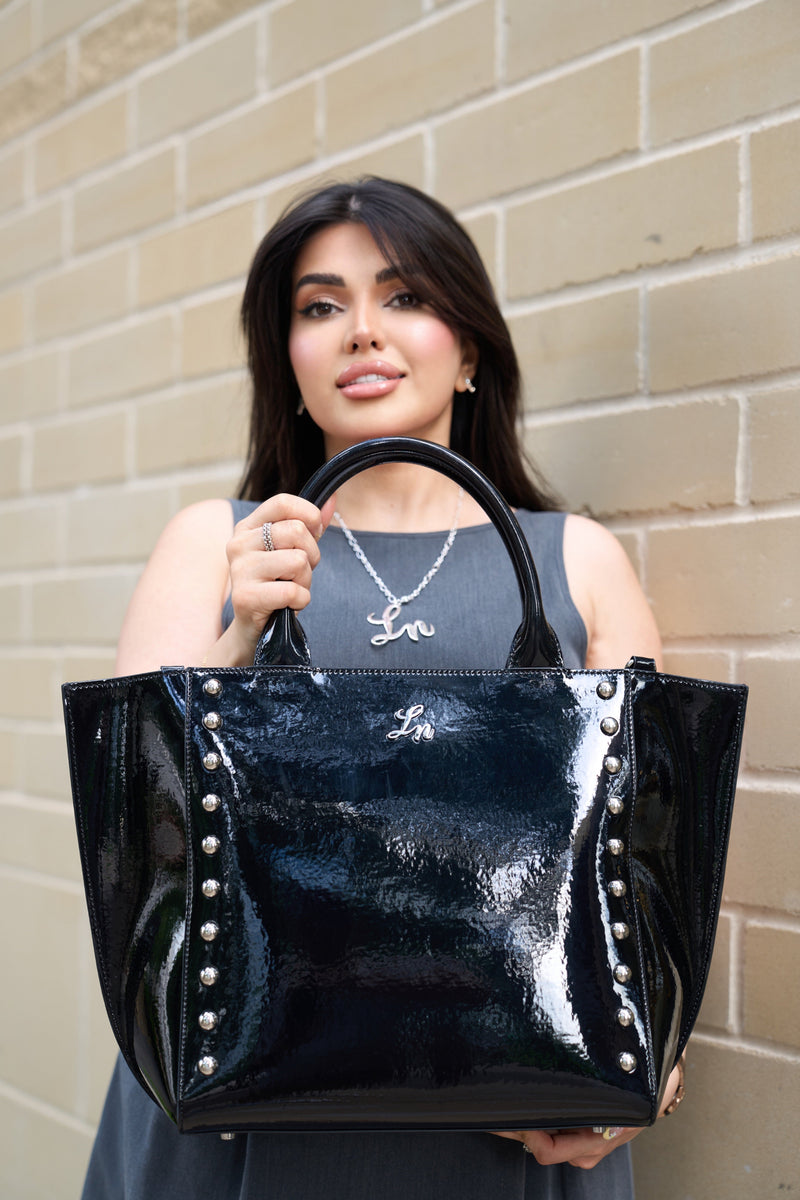Woman holding a black patent leather handbag with studded details against a beige brick wall.