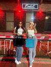 Two women posing in stylish outfits at a trendy outdoor venue, showcasing their bags and sunglasses.